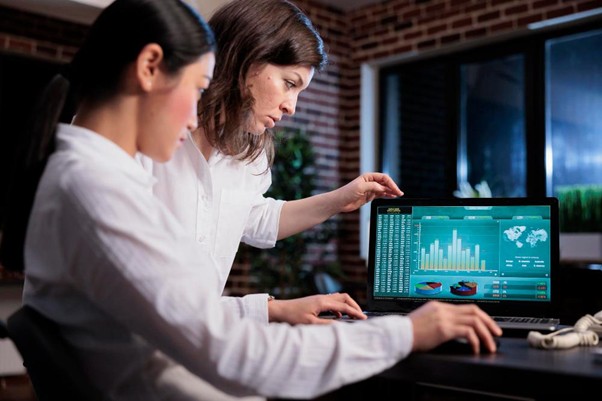 Two professional women analyzing complex data charts and performance metrics on a laptop screen in a modern office setting.